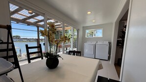 Ein Zimmer mit Tisch, Stühlen, einer Zimmerpflanze, einer Waschmaschine und Blick auf das Wasser.