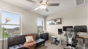A room with a sofa, a desk with two monitors, a ceiling fan, and large windows. - 3BR Sanctuary: Clean, Cozy & Complete with Pools and 20 min to DT Austin (Mustang Ridge)