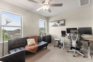A room with a sofa, a desk with two monitors, a ceiling fan, and large windows. - 3BR Sanctuary: Clean, Cozy & Complete with Pools and 20 min to DT Austin (Mustang Ridge)