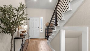 A hallway with a wooden floor, a white door, a staircase with white railings, and a plant on a console table. - 3BR Sanctuary: Clean, Cozy & Complete with Pools and 20 min to DT Austin (Mustang Ridge)