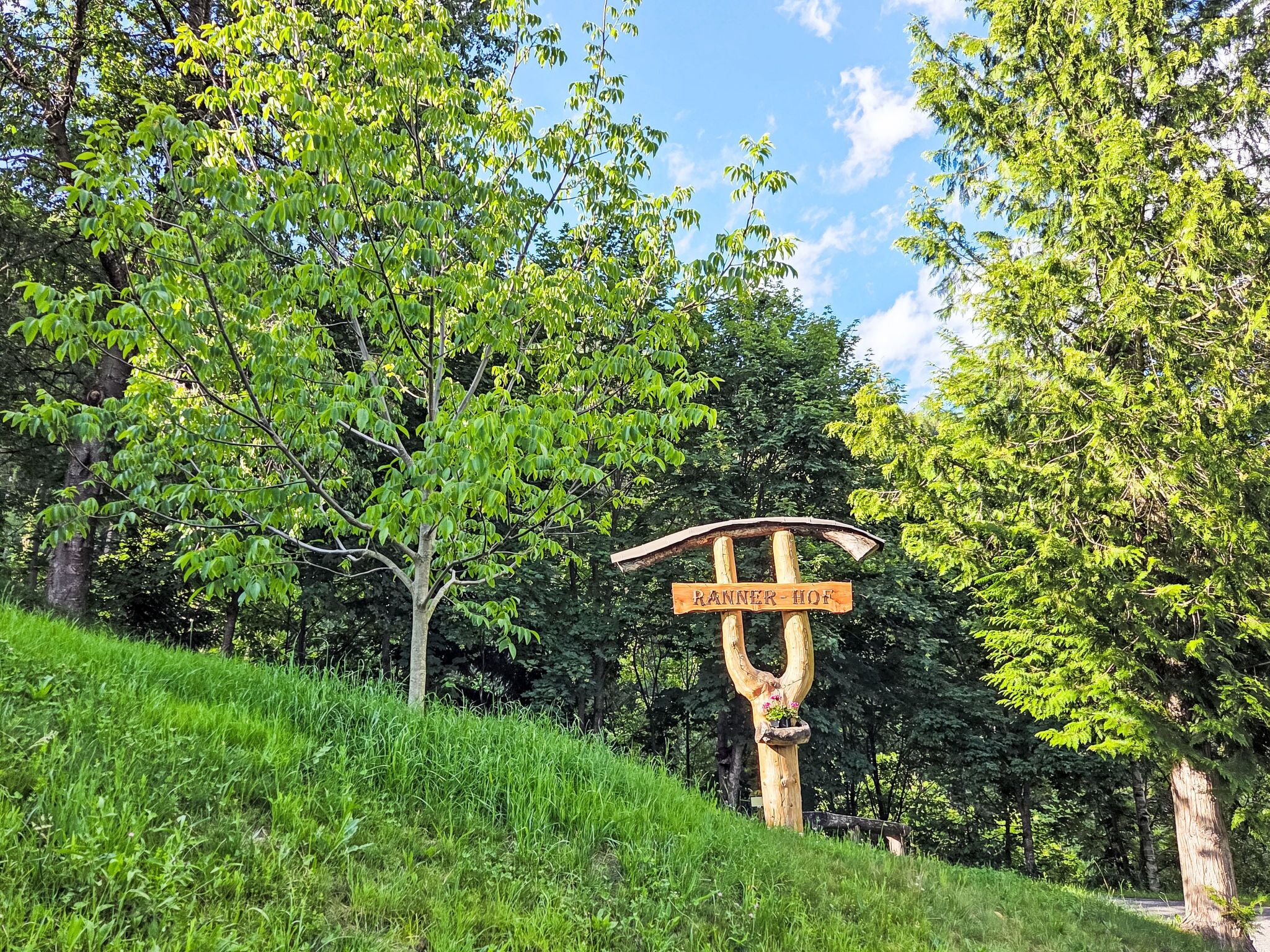 A wooden signpost with directional arrows and the text 'RANNER-HOF' in a natural setting with trees and grass.