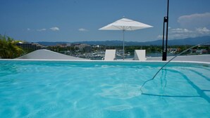 Uma piscina com água cristalina, um guarda-sol branco e vista para uma marina e montanhas ao fundo.