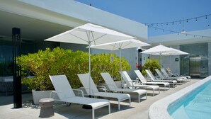 Área de piscina com cadeiras de sol brancas e guarda-sóis, cercada por vegetação e fachada de um prédio moderno.