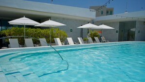 Piscina com espreguiçadeiras e guarda-sóis, emoldurada por um edifício moderno com fachada de vidro.