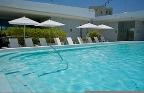 Ein Swimmingpool mit Liegestühlen und Sonnenschirmen, umgeben von einem modernen Gebäude mit Glasfassade.