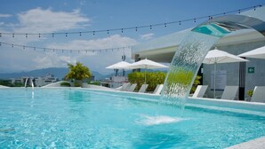 Uma piscina com um recurso aquático, cadeiras de descanso, guarda-sóis e vista para as montanhas ao fundo.