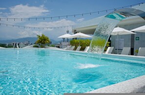 Ein Schwimmbecken mit Wasserspielen, Liegestühlen, Sonnenschirmen und Blick auf die Berge im Hintergrund.