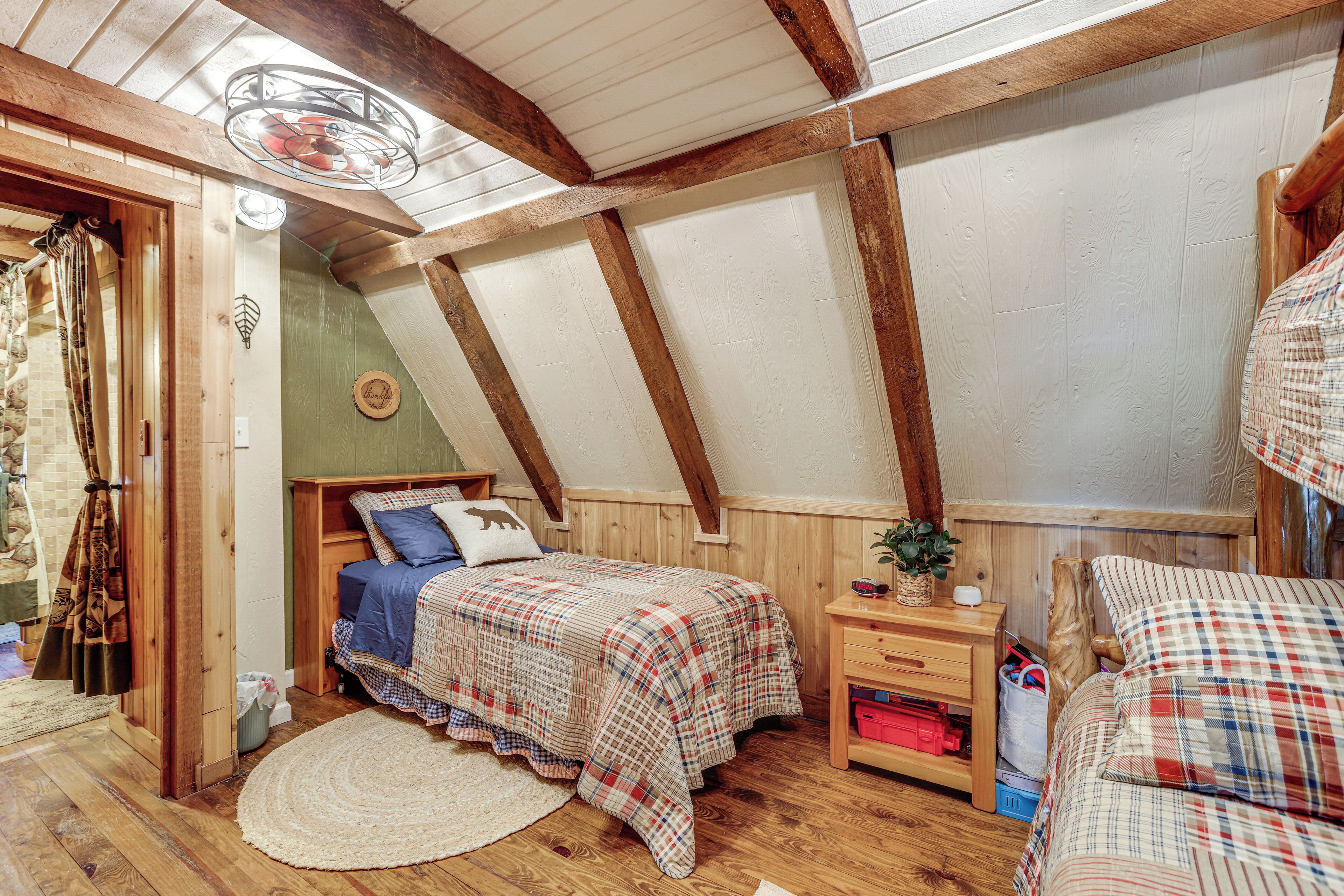 Une chambre mansardée chaleureuse avec un plafond en pente, des poutres en bois, un lit avec une couette à carreaux, une table de chevet, une petite table avec une plante et une fenêtre avec des rideaux.