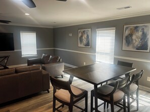 A modern living room with a dark brown sofa, a dining table with four chairs, and two large windows with blinds. - Elevated three master suites with jet tub, large kitchen, full of amenities (Camden)