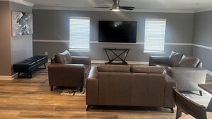 A modern living room with a ceiling fan, a flat-screen TV, and two windows with blinds. - Elevated three master suites with jet tub, large kitchen, full of amenities (Camden)