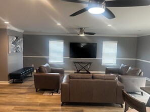 A modern living room with a ceiling fan, a flat-screen TV, and two windows with blinds. - Elevated three master suites with jet tub, large kitchen, full of amenities (Camden)