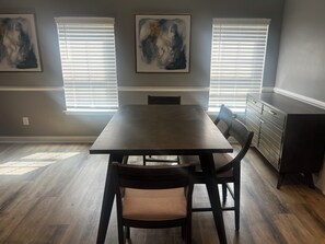 A dining area with a rectangular table, two chairs, and a bench. There are two windows with blinds, a wall-mounted painting, and a cabinet to the right. - Elevated three master suites with jet tub, large kitchen, full of amenities (Camden)