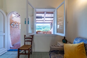 Ein Zimmer mit Fenster, ein Holzstuhl, eine Couch und Blick auf eine gebirgige Landschaft.