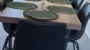 Et spisebord med grønne servietringe, sorte stole, en vase med blomster og udsigt til en have med hegn og træ.