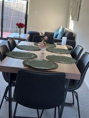 A dining table with green placemats, black chairs, a vase with flowers, and a view of a backyard with a fence and a tree. - New large 3BR,3bathrms townhouse in Gunghalin, mountain views,central location (Gungahlin)