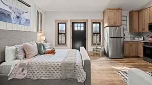 Un dormitorio moderno con una cama grande, una mesita de noche y un aparador. Hay una zona de cocina con armarios de madera y un refrigerador.