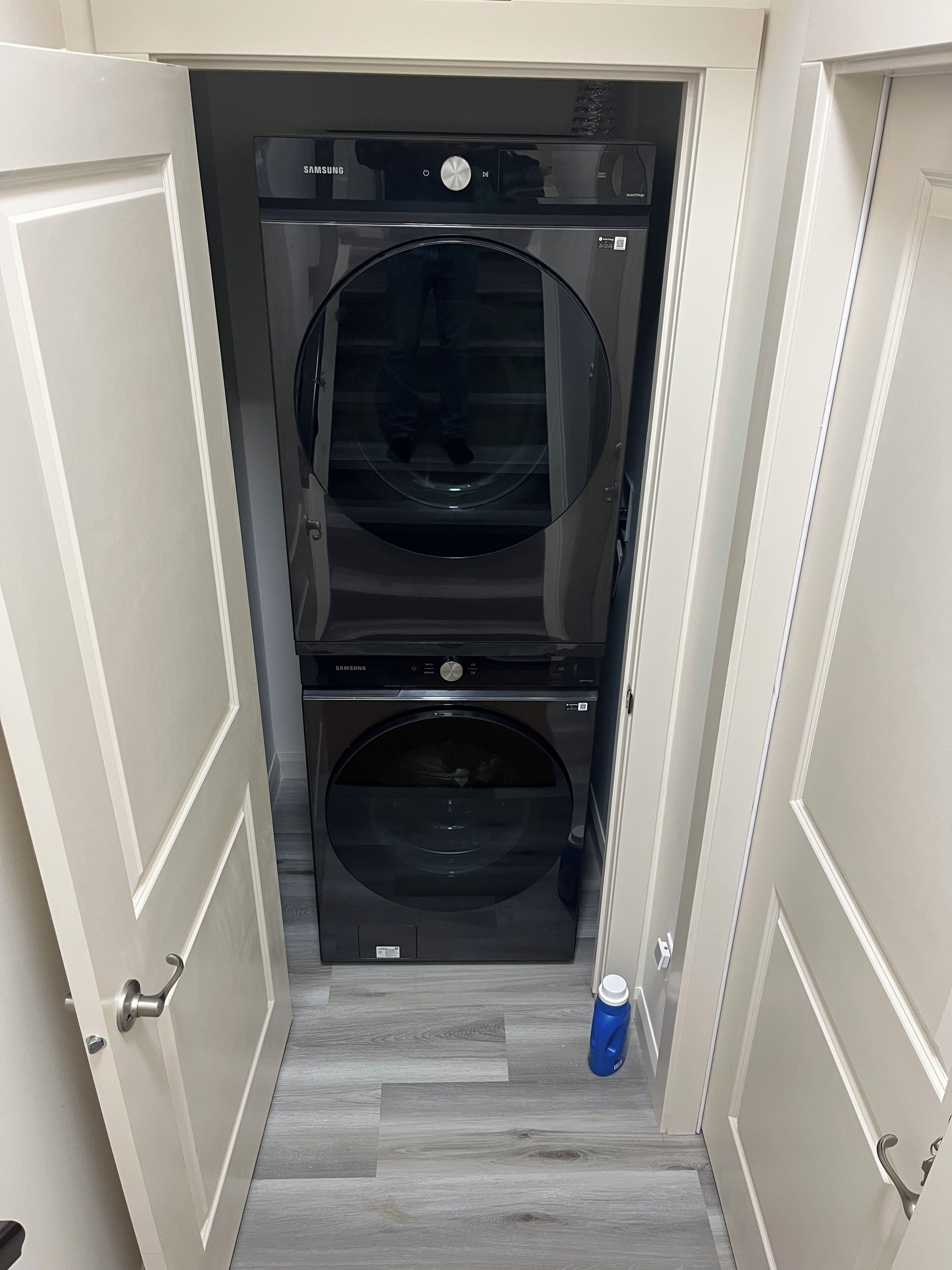 Une petite buanderie équipée de deux machines à laver Samsung, une bouteille d’eau bleue posée sur le sol et une porte donnant sur une autre pièce.
