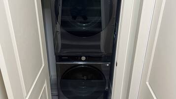 Une petite buanderie équipée de deux machines à laver Samsung, une bouteille d’eau bleue posée sur le sol et une porte donnant sur une autre pièce.