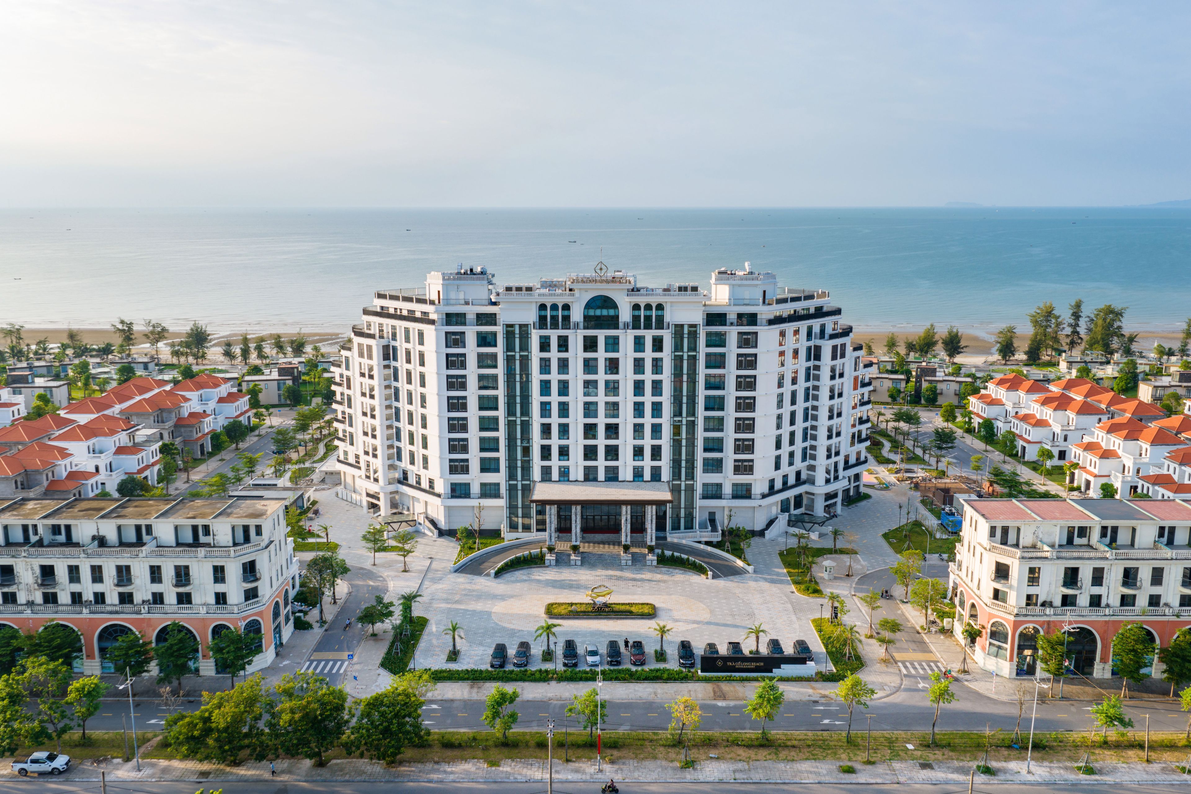 A multi-story hotel with a central courtyard, surrounded by residential buildings, overlooking a beach.