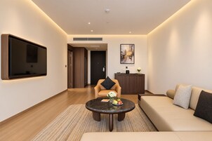 A modern living room with a flat-screen TV mounted on the wall, a round coffee table with fruits, a sofa, and a chair with a cushion.