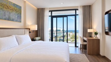 A hotel room with a large bed, a desk, a chair, a TV, and a balcony with a view.