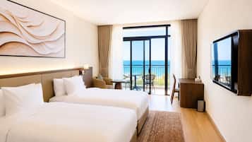 A hotel room with two beds, a desk, a chair, a TV, and a balcony with a view of the sea.