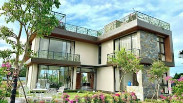 A modern two-story house with a balcony, large windows, and a garden with pink flowers.