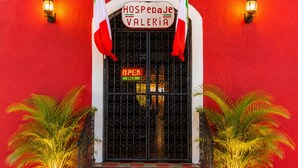 L’entrée d’un hôtel, avec un mur rouge, une enseigne indiquant « HOSPEDALJE VALERIA » et deux drapeaux.