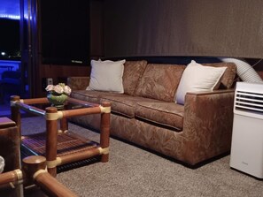 Um quarto com um sofá marrom estampado, uma pequena mesa de centro de madeira, um vaso com flores e um ar condicionado branco.