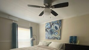 Un dormitorio con cama, ventilador de techo, ventana con cortinas y una obra de arte en la pared.