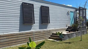 Una casa móvil de un nivel con techo plano, dos ventanas con contraventanas, una puerta azul y un pequeño jardín delantero con una planta en maceta.