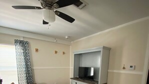 Una sala de estar con ventilador de techo, televisor sobre un mueble, sofá marrón, alfombra estampada y ventana con persianas.