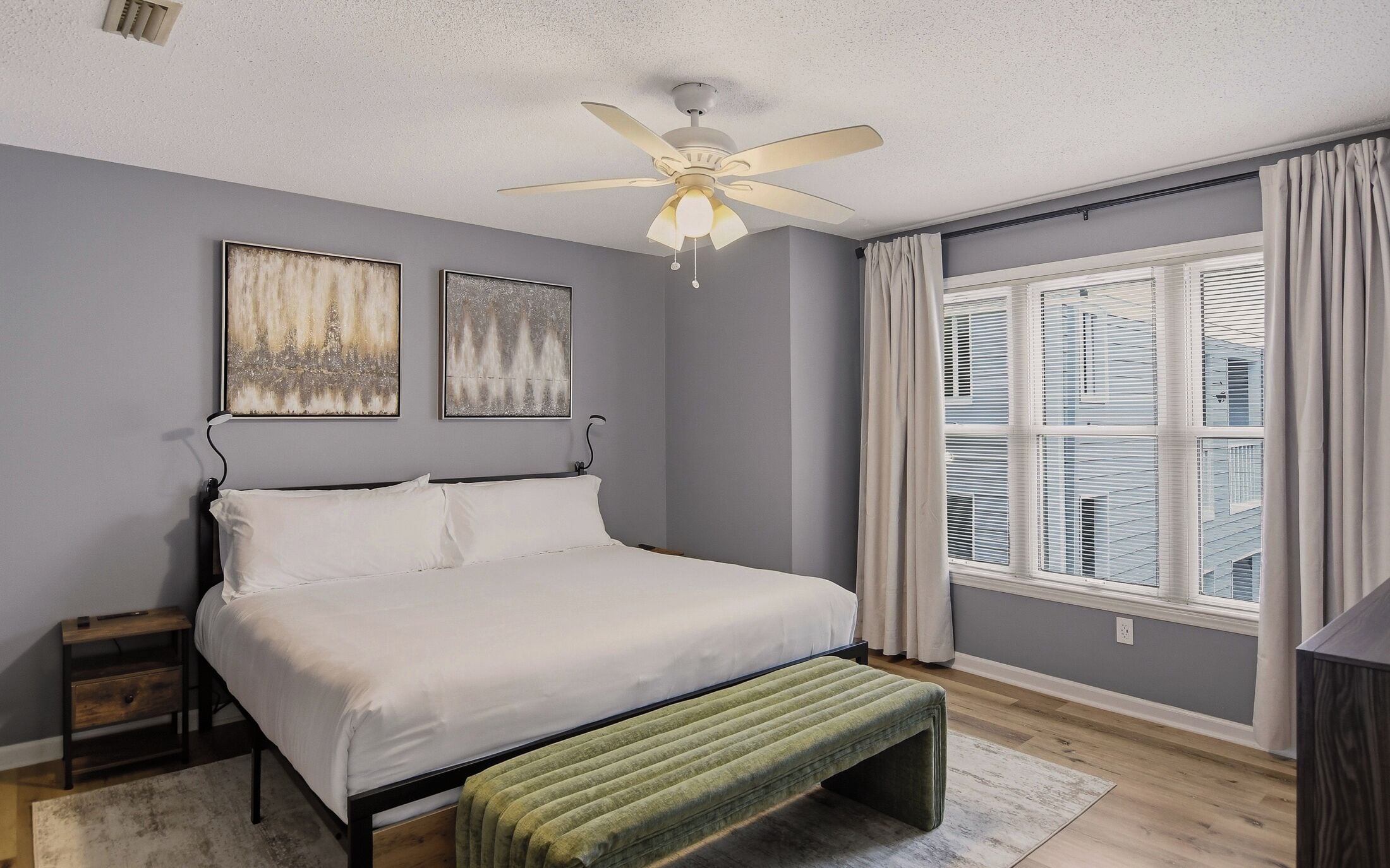 A bedroom with a bed, a nightstand, a bedside table, a ceiling fan, and a window with blinds.