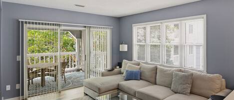 A living room with a sectional sofa, a glass coffee table, and a view through a sliding glass door to an outdoor area with a patio table and chairs.