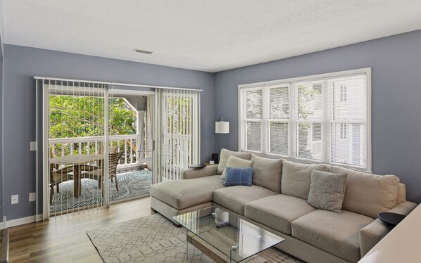 A living room with a sectional sofa, a glass coffee table, and a view through a sliding glass door to an outdoor area with a patio table and chairs.