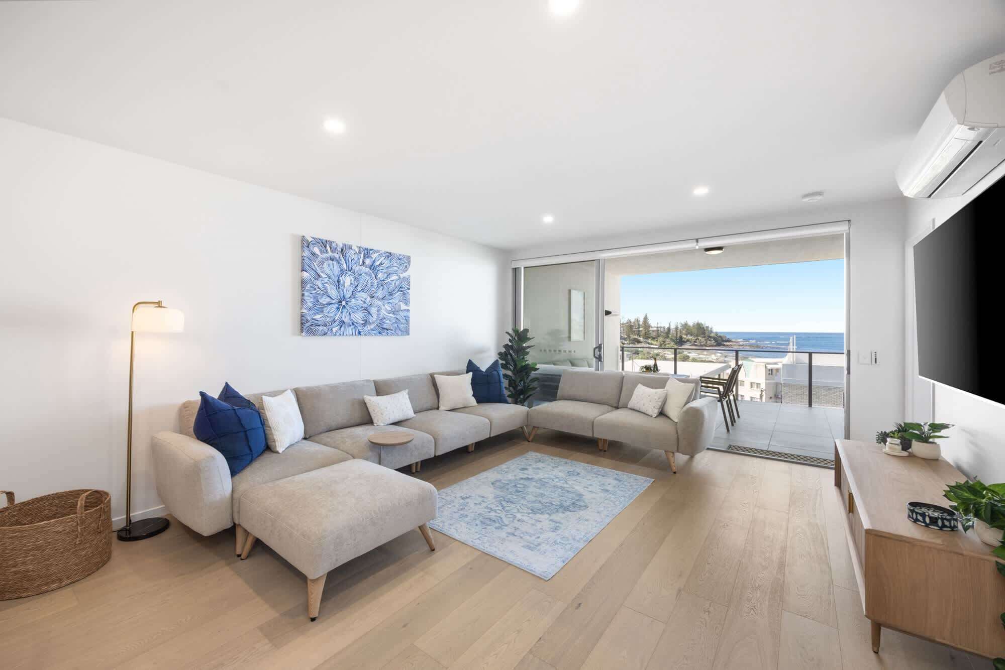Un salon moderne avec un canapé d’angle, une lampe sur pied, une grande œuvre d’art et une vue sur la mer depuis un balcon.