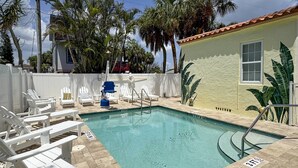 Uma piscina com cadeiras de descanso brancas e uma cadeira azul, cercada por palmeiras e um edifício com telhado vermelho.