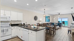 A spacious kitchen with white cabinetry, a central island, and a dining area with wooden furniture. - Emerald Dunes 502•Updated•Beachfront•Pool +Hot Tub•Beach Gear•Nearby Dining (Destin)