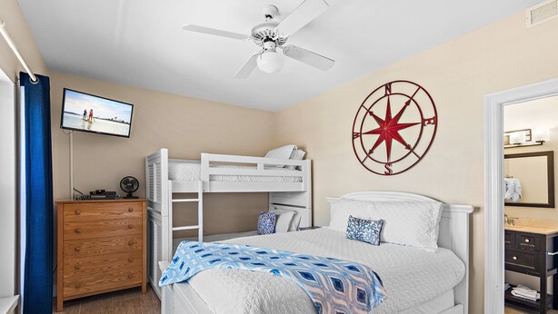 A bedroom with a bunk bed, a ceiling fan, a wall clock, a television, and a bathroom with a mirror and sink. - Emerald Dunes 502•Updated•Beachfront•Pool +Hot Tub•Beach Gear•Nearby Dining (Destin)
