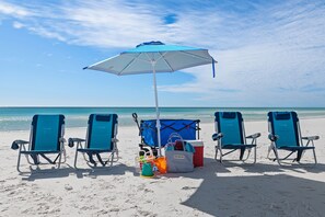 Eine Strandansicht mit vier blauen Liegestühlen, einem blau-weißen Sonnenschirm, einer Kühlbox und Strandspielzeug.