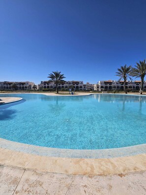 A large outdoor swimming pool with a clear blue water, surrounded by a tiled deck and palm trees. - Beachfront apartment with a terrace and ocean view (Marina Saidia)