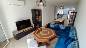 A modern living room with a flat-screen TV, a black entertainment unit, a blue sofa with patterned cushions, a wooden coffee table, and a dining area in the background. - Beachfront apartment with a terrace and ocean view (Marina Saidia)