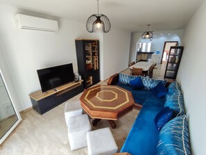 A modern living room with a flat-screen TV, a black entertainment unit, a blue sofa with patterned cushions, a wooden coffee table, and a dining area in the background. - Beachfront apartment with a terrace and ocean view (Marina Saidia)