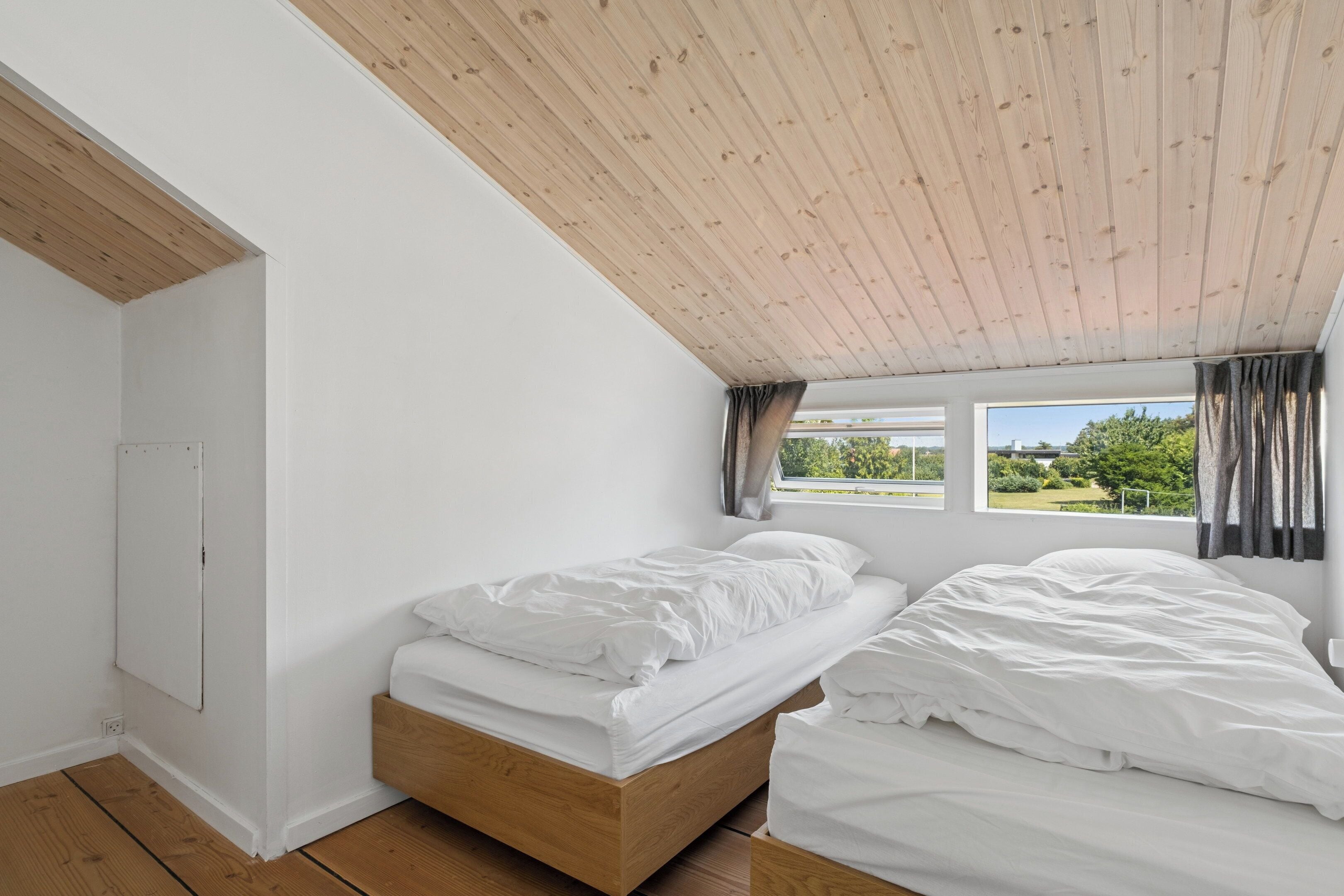 A bedroom with a wooden ceiling, a bed, a window with curtains, and a view of greenery outside.
