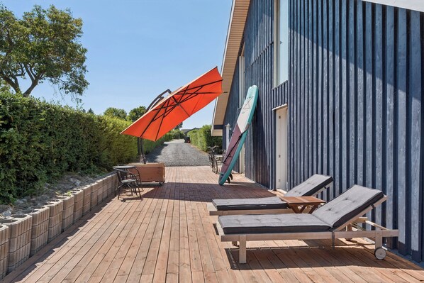 Una terrassa de fusta amb gandules i una taula, un parasol vermell i una taula de surf recolzada contra un edifici.
