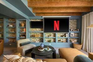 A cozy living room with a large flat-screen TV mounted on the wall, surrounded by shelves with decorative items and books. There are comfortable seating areas with sofas and a coffee table.