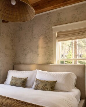 A bedroom with a bed, two pillows, a woven light fixture, a window with a view of trees, and a wall with a nature-themed pattern.