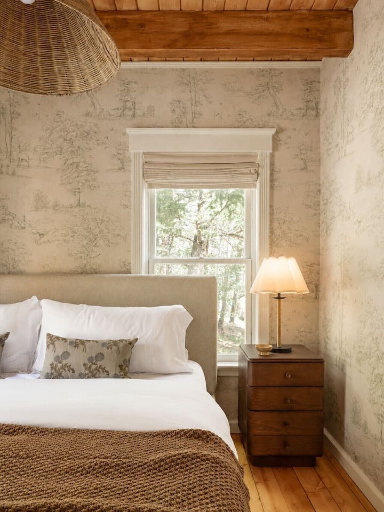 A bedroom with a bed, a nightstand, a lamp, and a window with a view of trees.