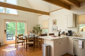 A modern kitchen with wooden beams, a dining area with chairs and a table, and a view of greenery outside.