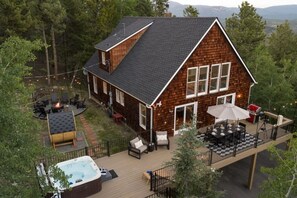 A two-story wooden house with a deck, outdoor seating, and a hot tub.